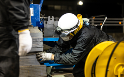 作業員がジャッキアップされた建物の基礎部分の隙間に、高さ調整用の鉄板を一枚ずつ手で慎重に挿入している様子の写真。